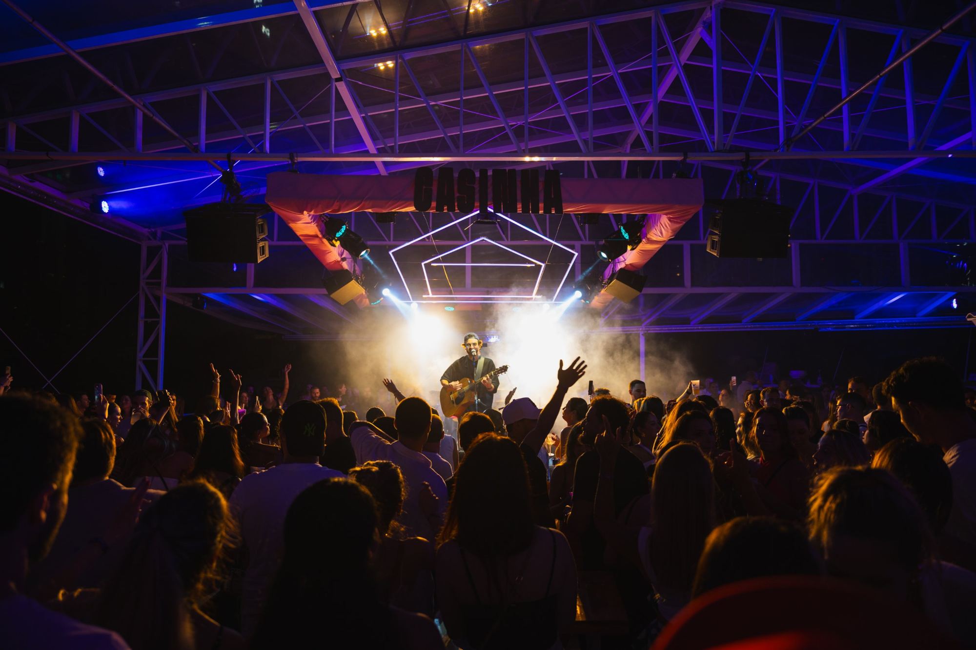 Simão no palco com voz e violão, público de mãos para o alto